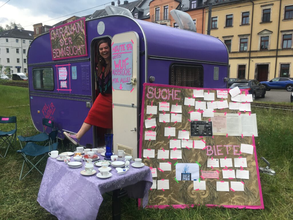 Der Caravan der Sehnsucht. Ein lila Wohnwagen mit einer Tafel mit der Aufschrift "SUCHE/BIETE" und einem Tisch mit Porzellanwaren darauf.